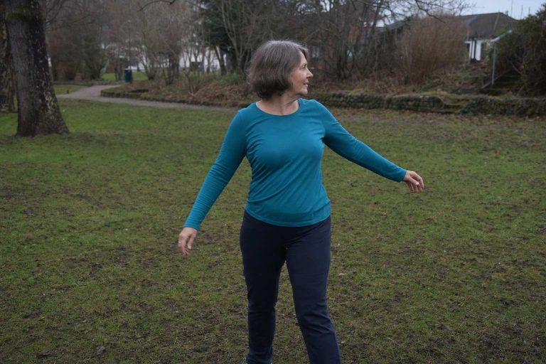 A woman in her sixties doing a gentle standing spine twist in a park in the morning